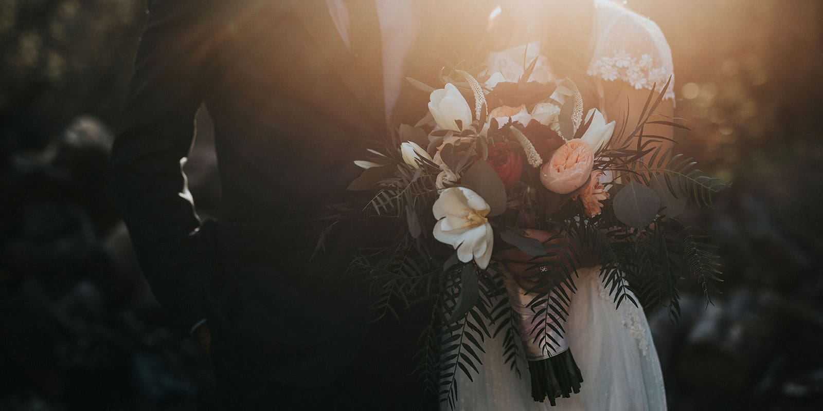 Due sposi davanti ad uno sfondo verde scuro; la sposa indossa un abito bianco e tiene in mano un mazzo di fiori; lo sposo indossa un abito nero.